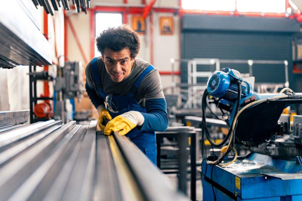 Ein Handwerker in blauer Arbeitskleidung und gelben Handschuhen arbeitet konzentriert an einem Metallstück in einer gut ausgestatteten Metallwerkstatt.