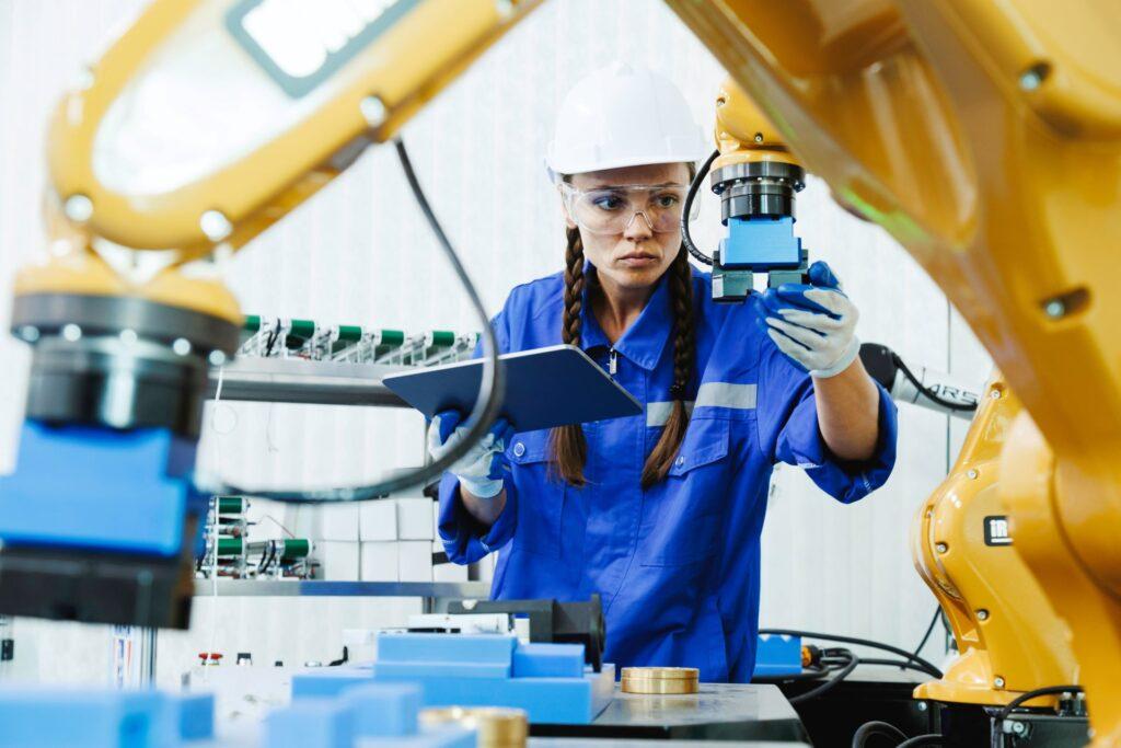 Eine Ingenieurin in blauer Arbeitskleidung und Schutzhelm führt eine Qualitätskontrolle an einem Werkstück mit Unterstützung einer Drehmaschine in einer modernen Produktionsumgebung durch.