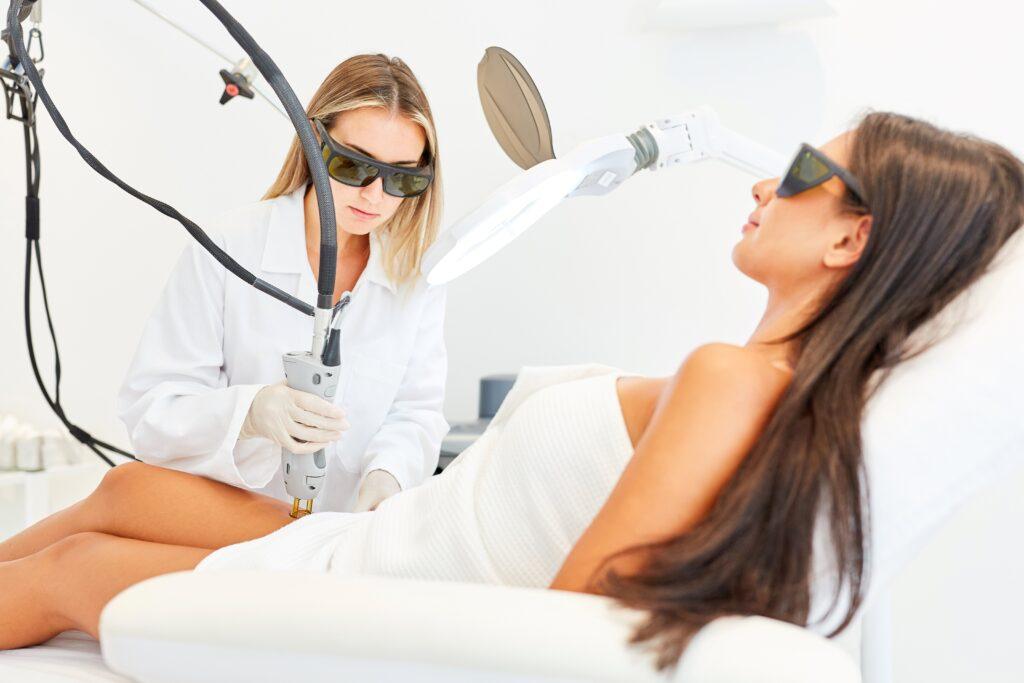 Eine Kosmetikerin führt eine Laserhaarentfernung an einer Frau durch, die auf einer Behandlungsliege liegt. Beide tragen Schutzbrillen.